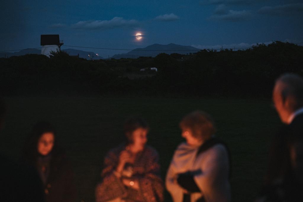 August wedding guests gather around a fire pit on Anglesey with the moon showing above Snowdonia, from an Anglesey Wedding Photographer